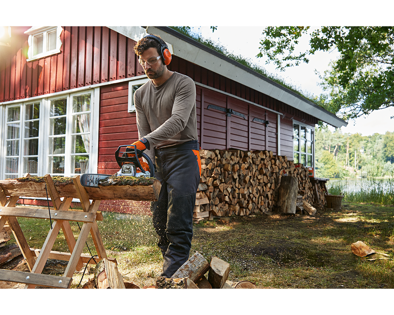 Man sawing a trunk into firewood with a STIHL petrol driven chainsaw MS 182 in front of a red wooden hut on the lakeshore