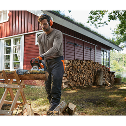 Man sawing a trunk into firewood with a STIHL petrol driven chainsaw MS 182 in front of a red wooden hut on the lakeshore